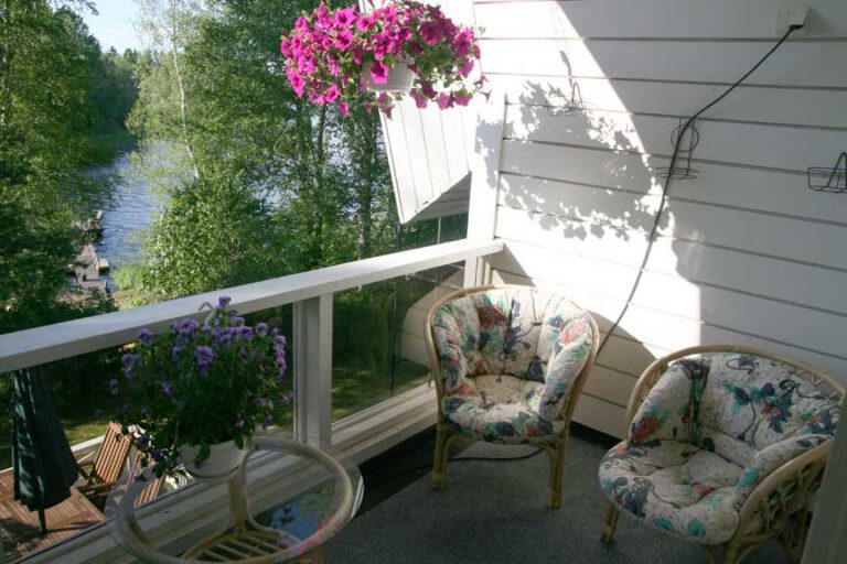 A covered balcony with wicker chairs and a hanging flower basket, the lake in the background.